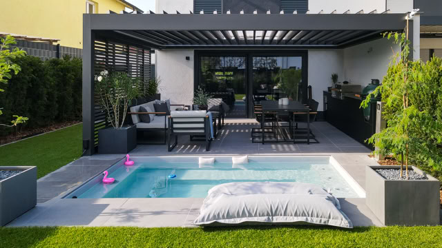 Moderne Terrasse mit Pergola, Sitzbereich, Esstisch und kleinem Pool im Vordergrund, umgeben von Rasen und Pflanzen.